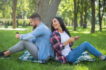 loving couple sitting on a plaid blanket in the park and use the phone, handsome guy and girl