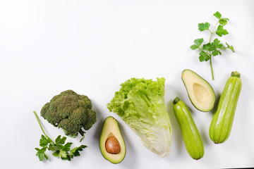 Bunch of greens and herbs laid on white countertop. Halved avocado, lettuce salad leaves, raw broccoli and other vegetables in row. Vegan diet concept. Close up, copy space, background, top view.