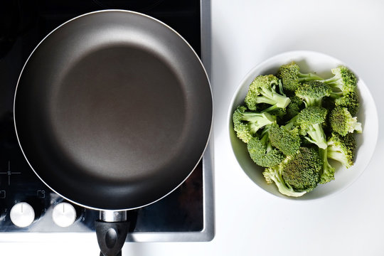 Pile of green raw uncooked broccoli in white ceramic bowl on countertop with empty frying pan on electric induction stove. Vegan diet concept. Close up, copy space, background, top view.