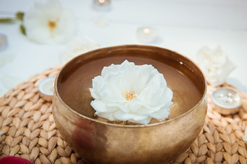 Close up Tibetan singing bowl with floating in water rose inside. Burning candles, flowers petals on the white wooden background. Meditation and Relax. Exotic massage. Copy space.