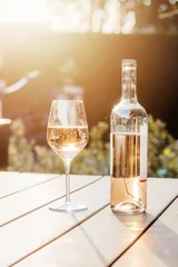 Poster Cold rose wine in bottle and glass on table in garden at sunset light. Sun reflecting in wine glass. Backyard outdoor summer lifestyle. © Tetiana Soares