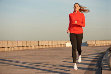 A jogging woman in red running shirt and black leggins on the street at sunrise. Running on concrete quay