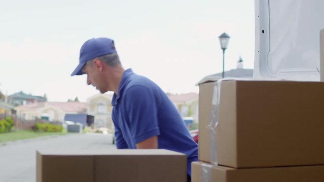 Handheld shot of male postal service worker in blue uniform loading packages into trunk of delivery van