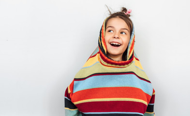 Closeup portrait of a happy little girl smiling broadly, playing with big colorful sweater on gray background. Smiling child peeks out of a sweater, looking up to the blank copy space over white wall