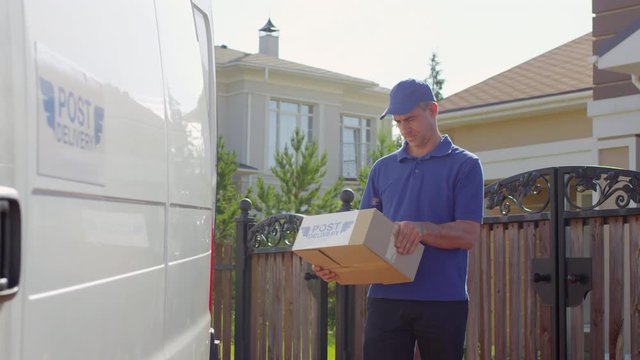 Handheld portrait shot of male courier in uniform checking information on package, then looking at camera and posing beside white delivery van