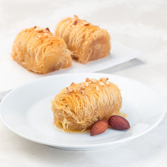 Greek pastry Kataifi with shredded filo dough stuffed with almond nuts, in honey syrup, on  white plate, square format