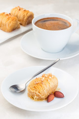 Greek pastry Kataifi with shredded filo dough stuffed with almond nuts, in honey syrup, on a white plate, served with cup of coffee, vertical