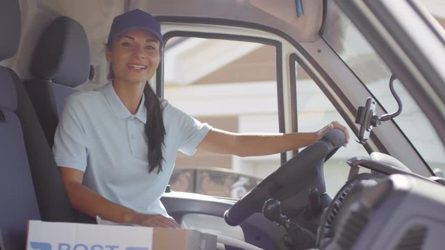 Handheld portrait shot of happy female postal service worker sitting in drivers seat of delivery van and talking while smiling and looking at camera