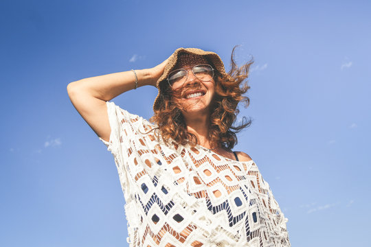 Trendy woman with hat enjoying summer holiday outdoor. Portrait of smiling girl with glasses. Wind blown in the hair and blue sky. Happy fashion female tourist. Tourism freedom and carefree concept.