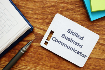 Text sign showing Skilled Business Communicator. The text is written on a small wooden board with exclamation mark silhouette. There are notebook, pen, colored papers on the photo.