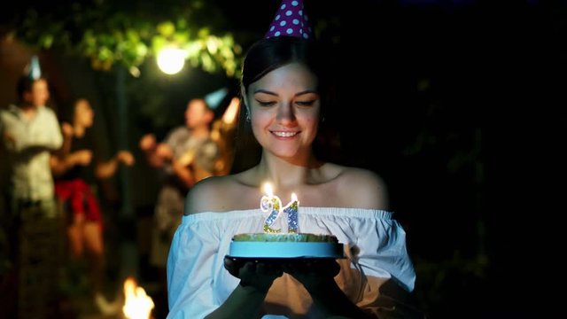 Beautiful woman stands with a cake and candles for her birthday, makes a wish, friends are dancing in the background, a party in a country house at night