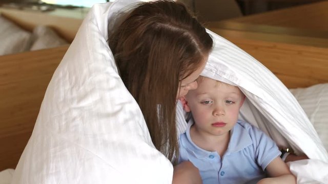 Mother hugs her little sad son on the bed under the blanket. Portrait of a sad baby on the bed, mom hugs and calms the child. Slow motion.