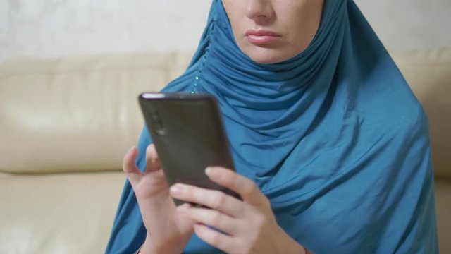 beautiful muslim woman in blue hijab using a mobile phone sitting on a sofa