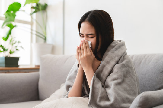 sick asian young woman sneezing into tissue paper while covered with a blanket and sitting on sofa in the living room