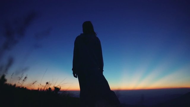 Amazed female wanderer standing on top of the hill at sunset observing nature panorama. Back view of excited woman enjoying scenery of mountain peaks after sunset.