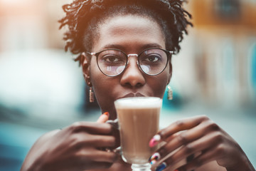 A close-up portrait of a young charming black woman in spectacles outdoors holding a glass of delicious coffee latte; an African girl in glasses and with nails art is drinking cocoa in a street cafe
