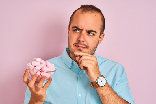 Young man holding bowl with marshmallows standing over isolated pink background serious face thinking about question, very confused idea