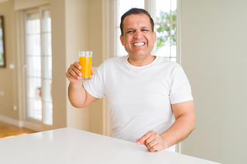 Middle age man drinking a glass of orange juice at home with a happy face standing and smiling with a confident smile showing teeth