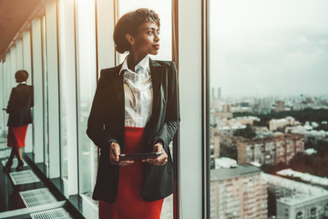 An African-American woman entrepreneur a digital tablet is leaning against a panoramic window of an office business skyscraper and looking in a distance; biracial businesswoman with a tablet pc - Powered by Adobe