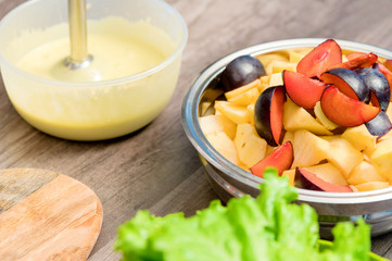 Close-up of whipping a mixture of homemade mayonnaise with a blender in a plastic bowl. Sliced apples and plums in a metal bowl near
