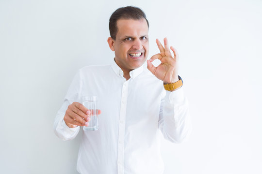 Middle age man drinking a glass of water over white background doing ok sign with fingers, excellent symbol