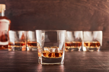 A glass of whiskey on a wooden table. In the background, four glasses of whiskey and a full bottle of whiskey.