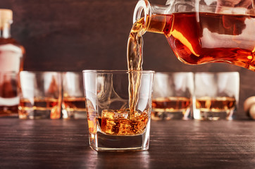 A glass of whiskey on a wooden table in which poured whiskey from a bottle. In the background are four glasses of whiskey, a full bottle of whiskey and a wooden bottle stopper.