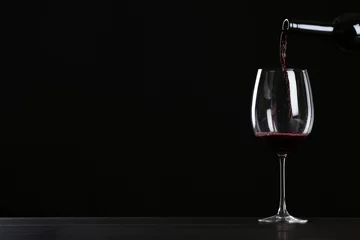 Gordijnen Pouring wine from bottle into glass on table against black background, space for text © New Africa