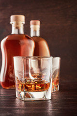 Two glasses of whisky with ice on a wooden table. In the background, two bottles of whiskey of different shapes.