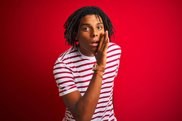 Young afro man with dreadlocks wearing striped t-shirt standing over isolated red background hand on mouth telling secret rumor, whispering malicious talk conversation