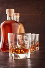 Three glasses of whiskey on the ice on the wooden table. In the background, two bottles of whiskey of different shapes.