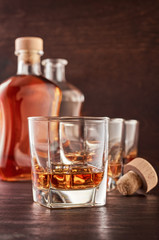 Three glasses of whiskey on the ice on the wooden table. In the background are two bottles of whiskey, full and open, as well as a wooden bottle stopper.