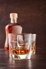 Two glasses of whisky with ice on a wooden table. There's a full bottle of whiskey in the background.