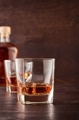 Two glasses of whisky with ice on a wooden table. There's a full bottle of whiskey in the background.