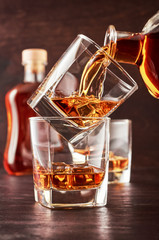 Two glasses of whisky with ice on a wooden table. One glass stands on the other. The glass is poured whiskey from the bottle. In the background is a full bottle of whiskey and a glass of whiskey.