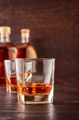 Two glasses of whisky with ice on a wooden table. In the background, two bottles of whiskey of different shapes.