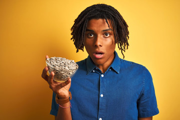 Afro american man with dreadlocks holding sunflowers seeds over isolated yellow background scared...