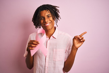 Afro american man with dreadlocks holding cancer ribbon over isolated pink background very happy pointing with hand and finger to the side