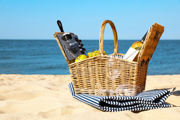 Wicker picnic basket with products and blanket on sunny beach. Space for text