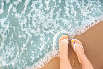 Top view of woman with white flip flops on sand near sea, space for text. Beach accessories - obrazy, fototapety, plakaty