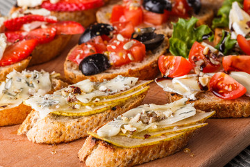 Assorted bruschetta with tomato, olives, camembert, blue cheese, lettuce, pear, honey on wooden board
