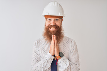 Young redhead irish architect man wearing security helmet over isolated white background praying with hands together asking for forgiveness smiling confident.