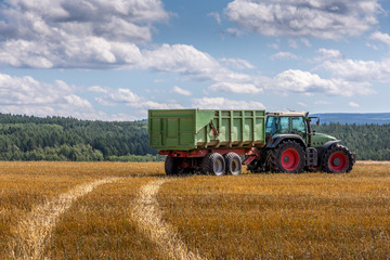 Tractor and trailer driving over a farm field leaving a tyre track in the dried grass on a cloudy blue sky day