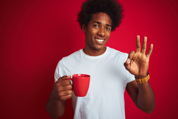 Young african american man drinking a cup of coffee standing over isolated red background doing ok sign with fingers, excellent symbol