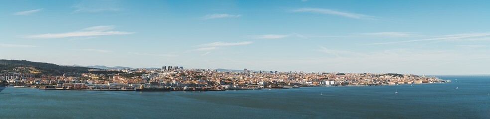 Fototapeta premium Panoramic view of Lisbon, Portugal from the Tagus river: waterline in the foreground, long river bank with a commercial docks near water, plenty of old houses and districts in the background