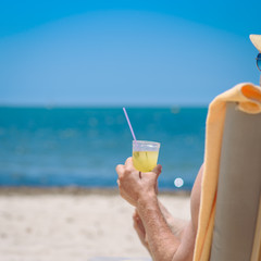 Man in sunhat is sitting on a sunbed at luxurious hotel near the ocean, he is enjoying his summer vacations and holding a glass of cocktail in his hand. Back view.