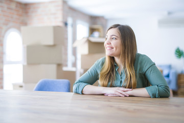 Young woman sitting on the table with cardboard boxes behind her moving to new home looking away to side with smile on face, natural expression. Laughing confident.