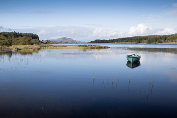 A tranquil inland Scottish loch has a rowing boat with reflection. Mountains and forestry on the horizon with blue summer sky - Image