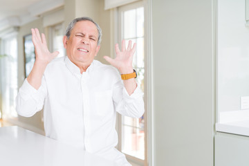 Fototapeta premium Handsome senior man at home crazy and mad shouting and yelling with aggressive expression and arms raised. Frustration concept.
