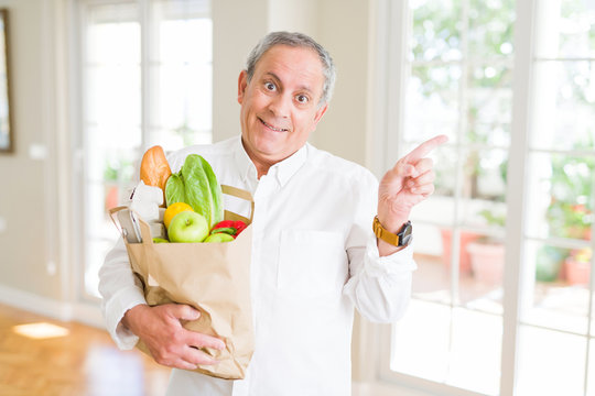 Handsome senior man holding a paper bag of fresh groceries from the supermarket very happy pointing with hand and finger to the side